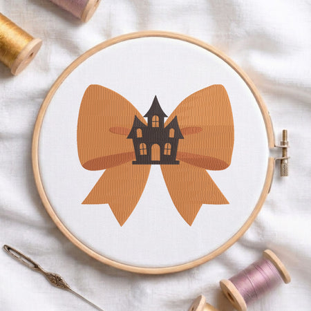 A white fabric stretched in a wooden embroidery hoop, featuring an embroidered design of a large orange bow centered with a spooky black haunted house.