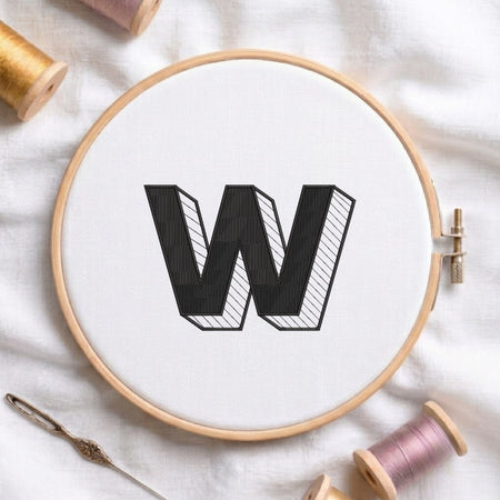 A wooden embroidery hoop holding white fabric with a large black outlined letter W stitched in the center. The hoop is styled on a white fabric background with embroidery thread spools and a needle nearby.