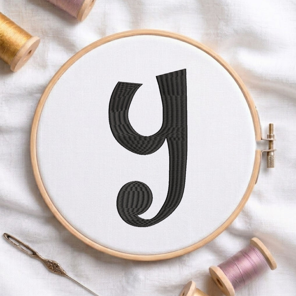 A wooden embroidery hoop holding white fabric with a large black decorative letter 'y' stitched in the center. The hoop is styled on a white fabric surface with spools of thread and a needle nearby.