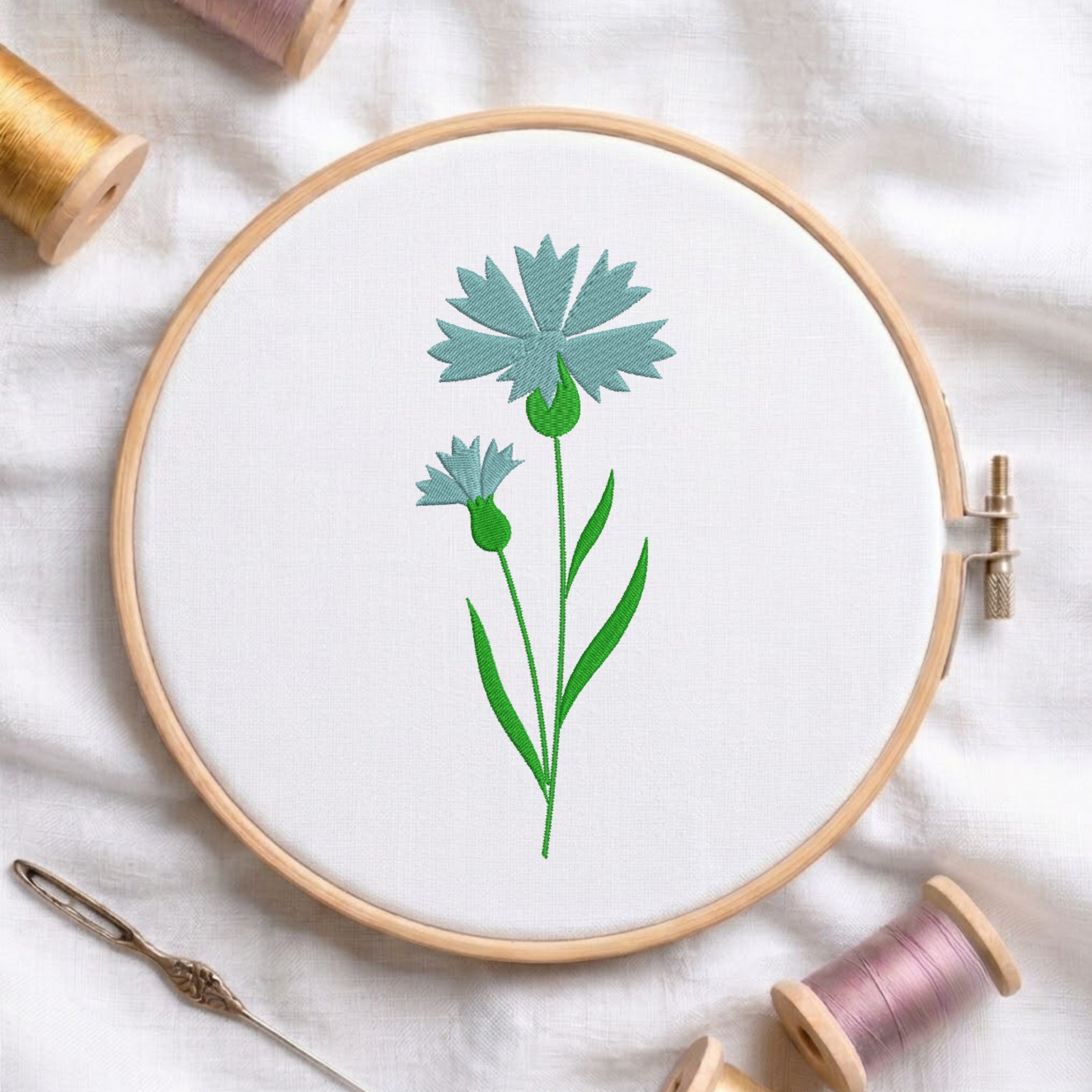 Soft blue cornflower wildflower with green stem embroidered inside a round wooden hoop with thread spools.