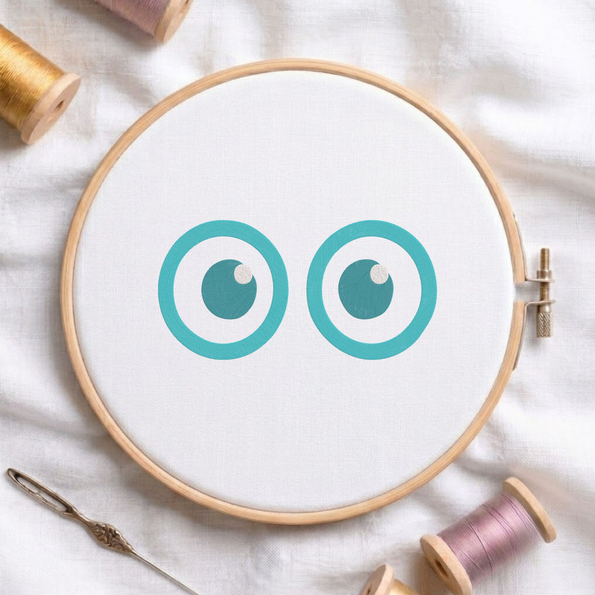 A wooden embroidery hoop holding white fabric with two teal cartoon eyes, surrounded by spools of thread and a needle on a white fabric background.