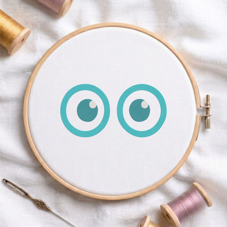 A wooden embroidery hoop holding white fabric with two teal cartoon eyes, surrounded by spools of thread and a needle on a white fabric background.