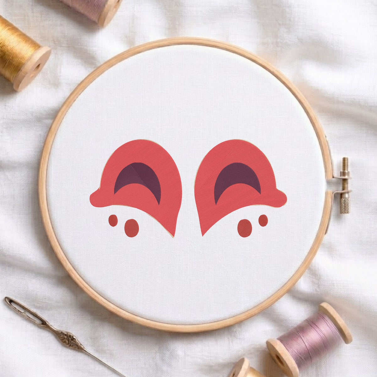Pinkish coral and dark purple animal ears embroidery pattern centered in a round wooden hoop with thread spools.