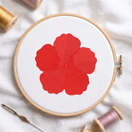 A wooden embroidery hoop holding white fabric with a large red flower design, surrounded by spools of thread and a needle on a white cloth background.