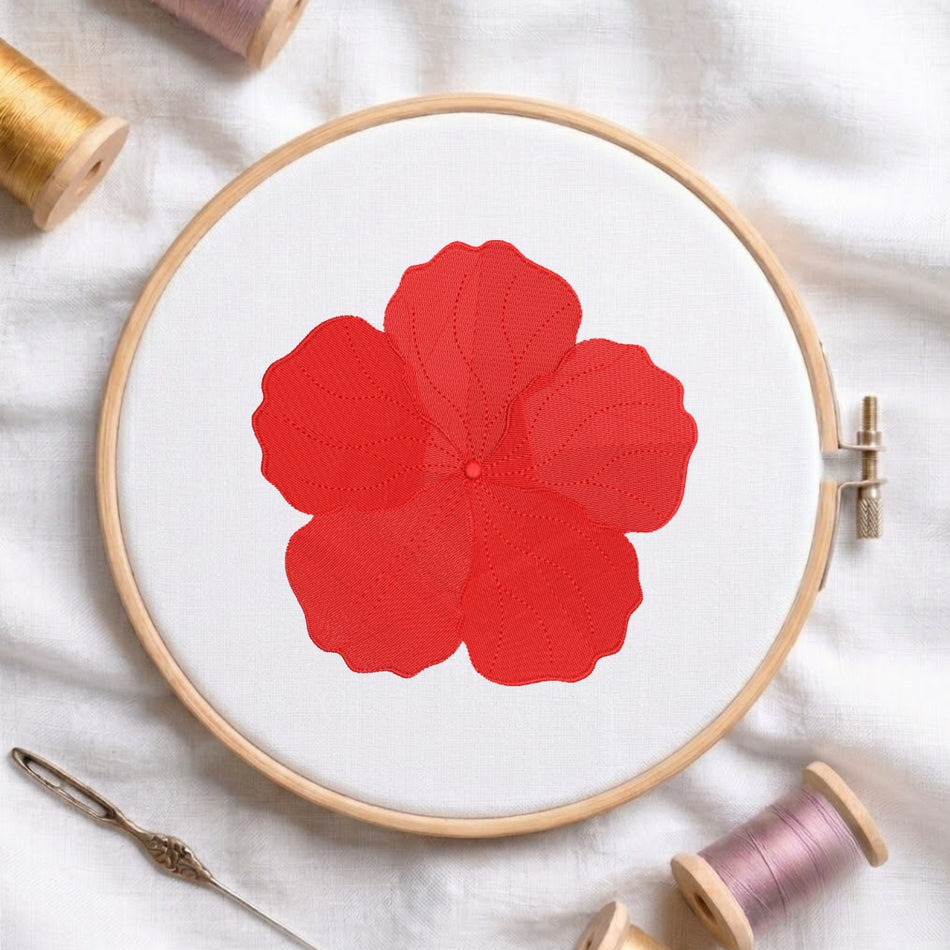 A wooden embroidery hoop holding white fabric with a large red flower design, surrounded by spools of thread and a needle on a white cloth background.
