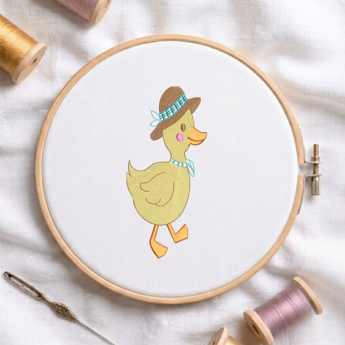 A circular embroidery hoop holding white fabric with a stitched duck design wearing a brown hat and blue bow tie, surrounded by spools of thread and a needle on a white fabric background.