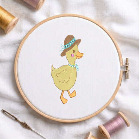 A circular embroidery hoop holding white fabric with a stitched duck design wearing a brown hat and blue bow tie, surrounded by spools of thread and a needle on a white fabric background.
