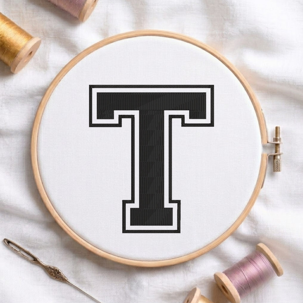 A wooden embroidery hoop holding white fabric with a large black letter T outlined in white. A needle lies at the lower left, and spools of thread are arranged around the hoop on a white fabric background.