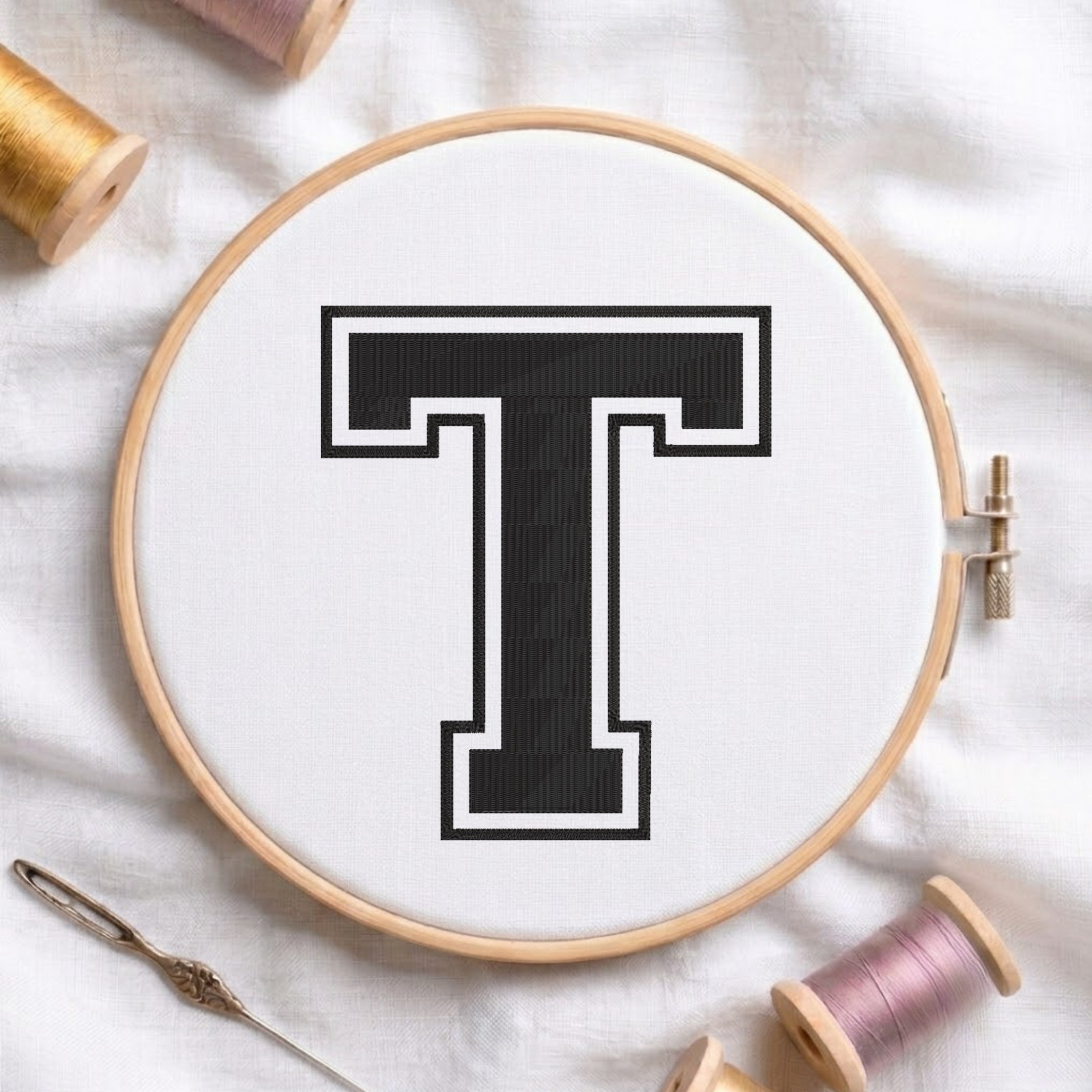 A wooden embroidery hoop holding white fabric with a large black letter T outlined in white. A needle lies at the lower left, and spools of thread are arranged around the hoop on a white fabric background.