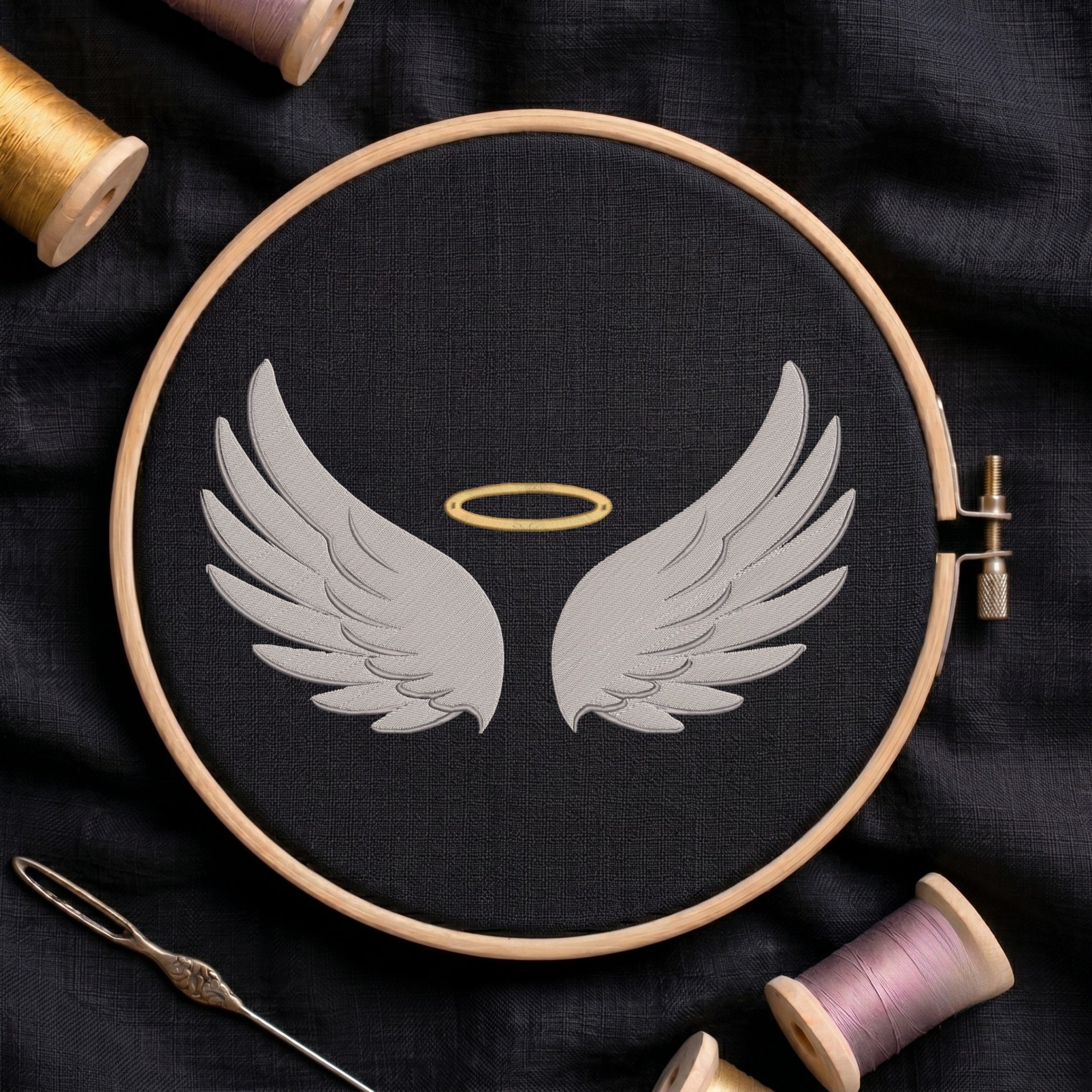 A circular embroidery hoop holding black fabric with a stitched design of two white angel wings and a small gold halo, surrounded by spools of thread and a needle on a dark fabric background.