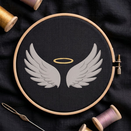 A circular embroidery hoop holding black fabric with a stitched design of two white angel wings and a small gold halo, surrounded by spools of thread and a needle on a dark fabric background.