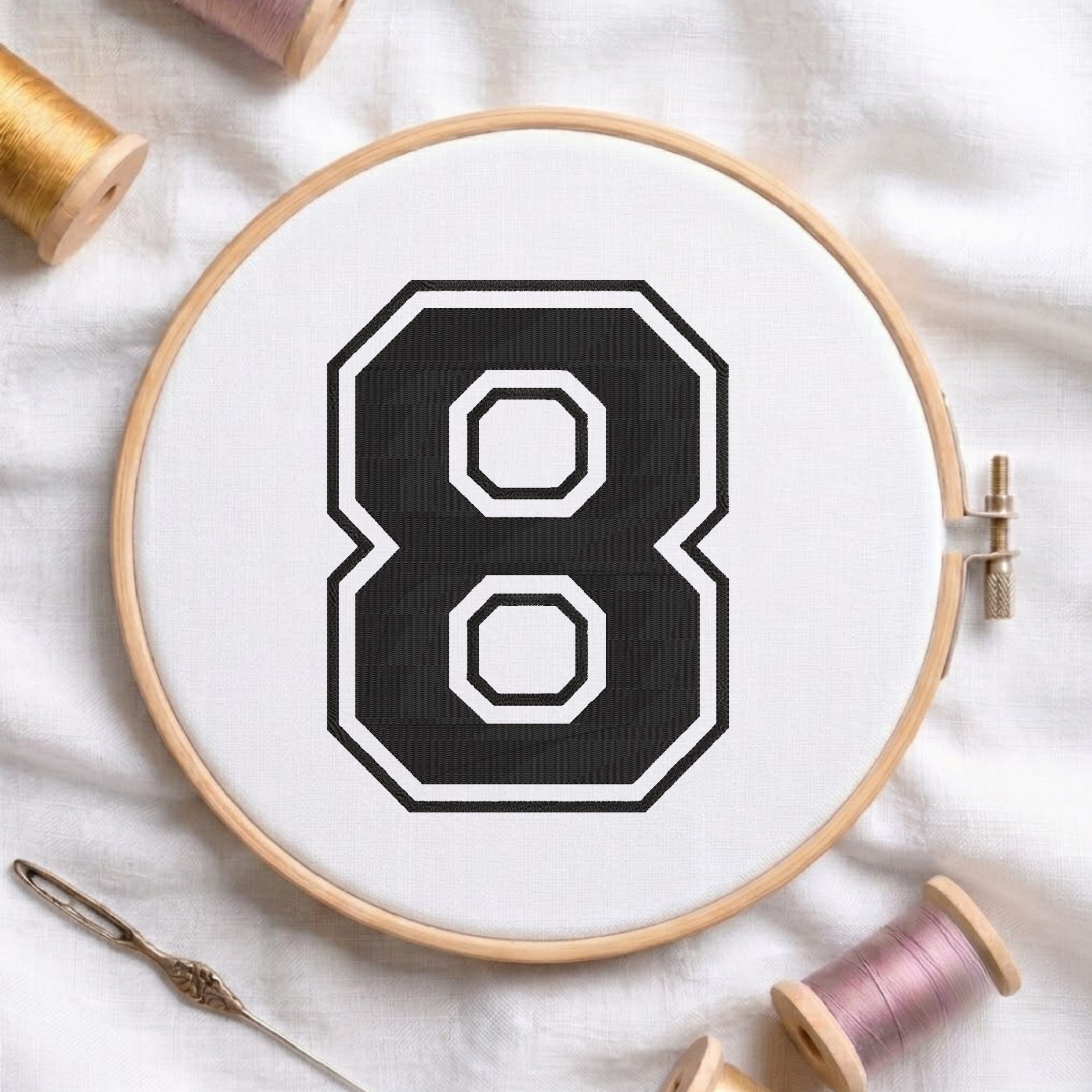 A wooden embroidery hoop holding white fabric with a large black number 8 design, surrounded by spools of thread and a needle on a white fabric background.