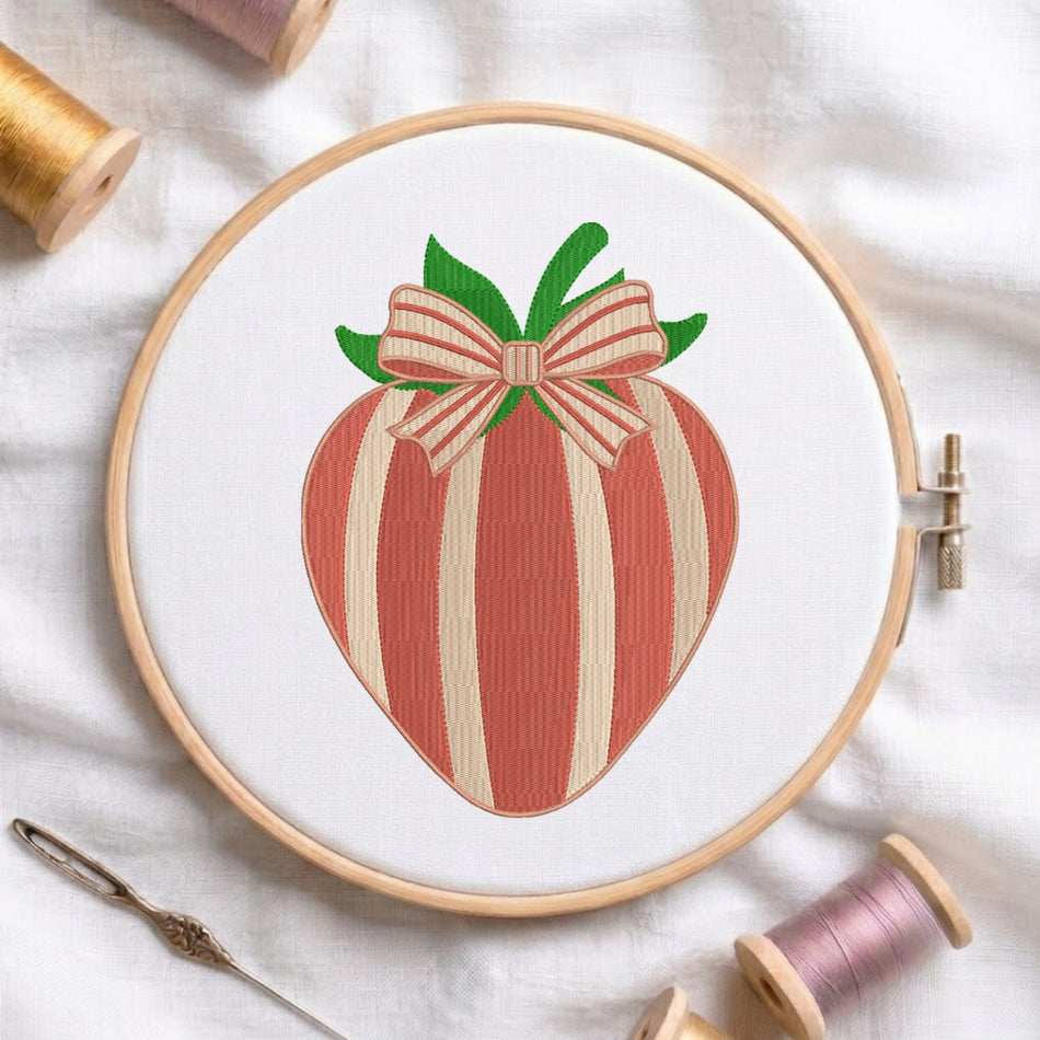 A wooden embroidery hoop holds white fabric with a stitched strawberry design in coral red and cream stripes, topped with green leaves and a matching striped bow. Spools of thread and a needle are arranged around it on a white fabric background.