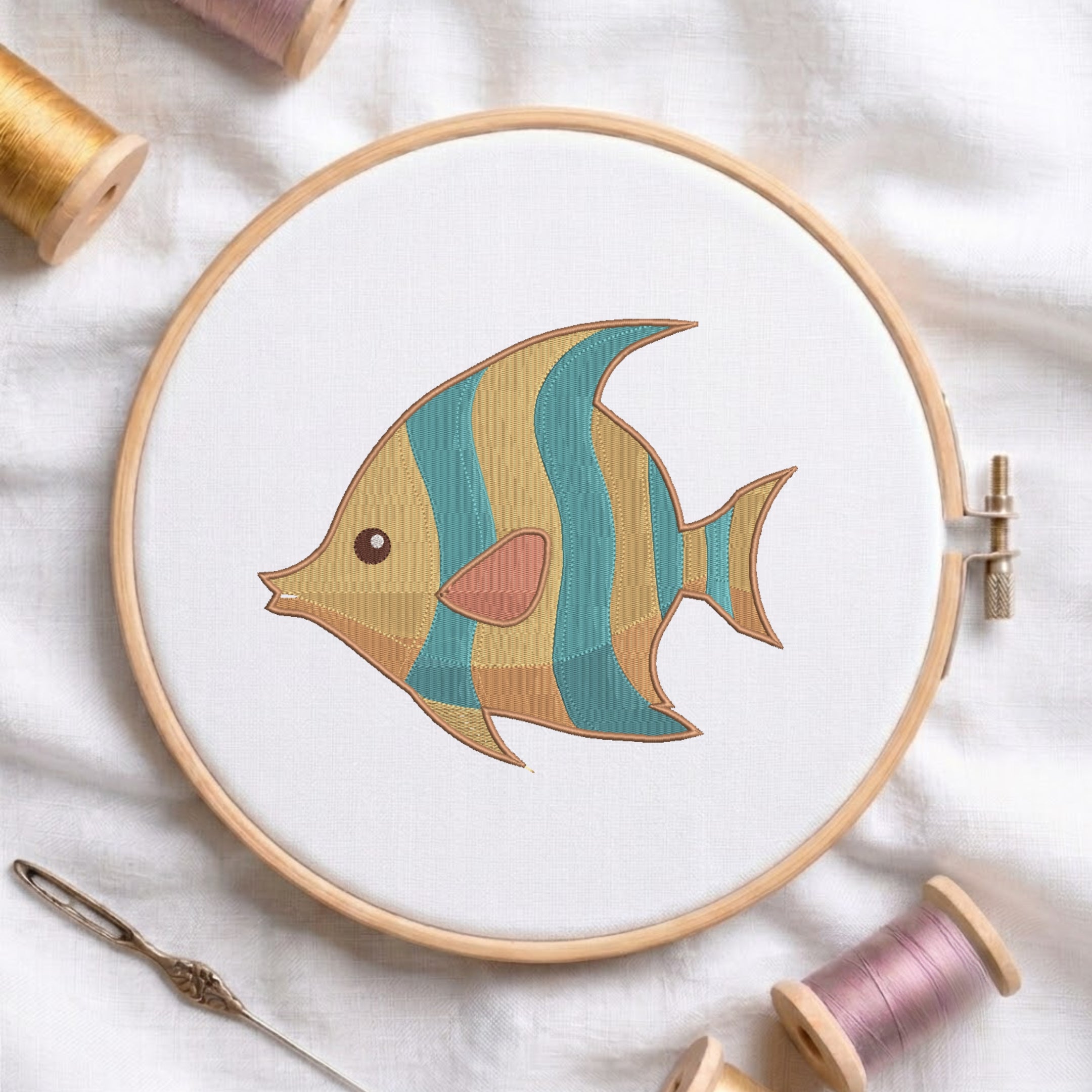 A wooden embroidery hoop holds white fabric with a stylized embroidered fish design in teal, mustard, peach, and brown. The hoop is styled on a white fabric background with spools of thread and a needle.