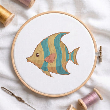 A wooden embroidery hoop holds white fabric with a stylized embroidered fish design in teal, mustard, peach, and brown. The hoop is styled on a white fabric background with spools of thread and a needle.