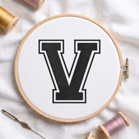 A wooden embroidery hoop holding white fabric with a large black letter V outlined in white. The hoop is styled on a white fabric surface with spools of thread and a needle around it.