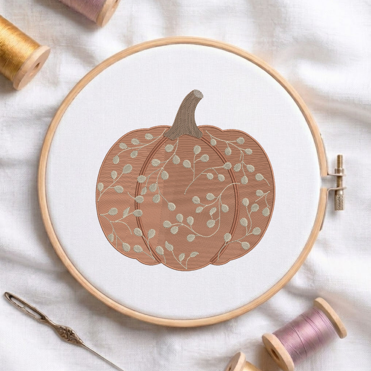 A circular embroidery hoop holding white fabric with a stitched pumpkin design in warm terracotta and pale sage green, surrounded by spools of thread and a needle on a white fabric background.