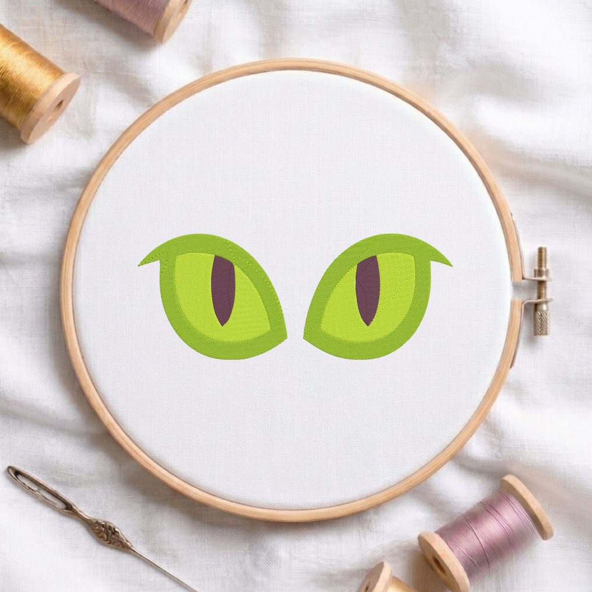 A circular embroidery hoop on white fabric, featuring a cartoon-style pair of green cat eyes with dark pupils. The hoop is styled with spools of thread, a needle, and a pen on a soft white fabric background.