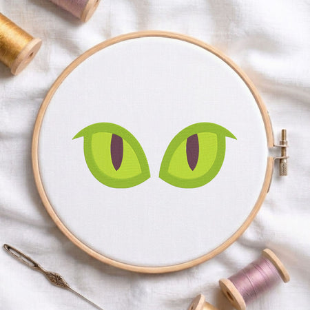 A circular embroidery hoop on white fabric, featuring a cartoon-style pair of green cat eyes with dark pupils. The hoop is styled with spools of thread, a needle, and a pen on a soft white fabric background.