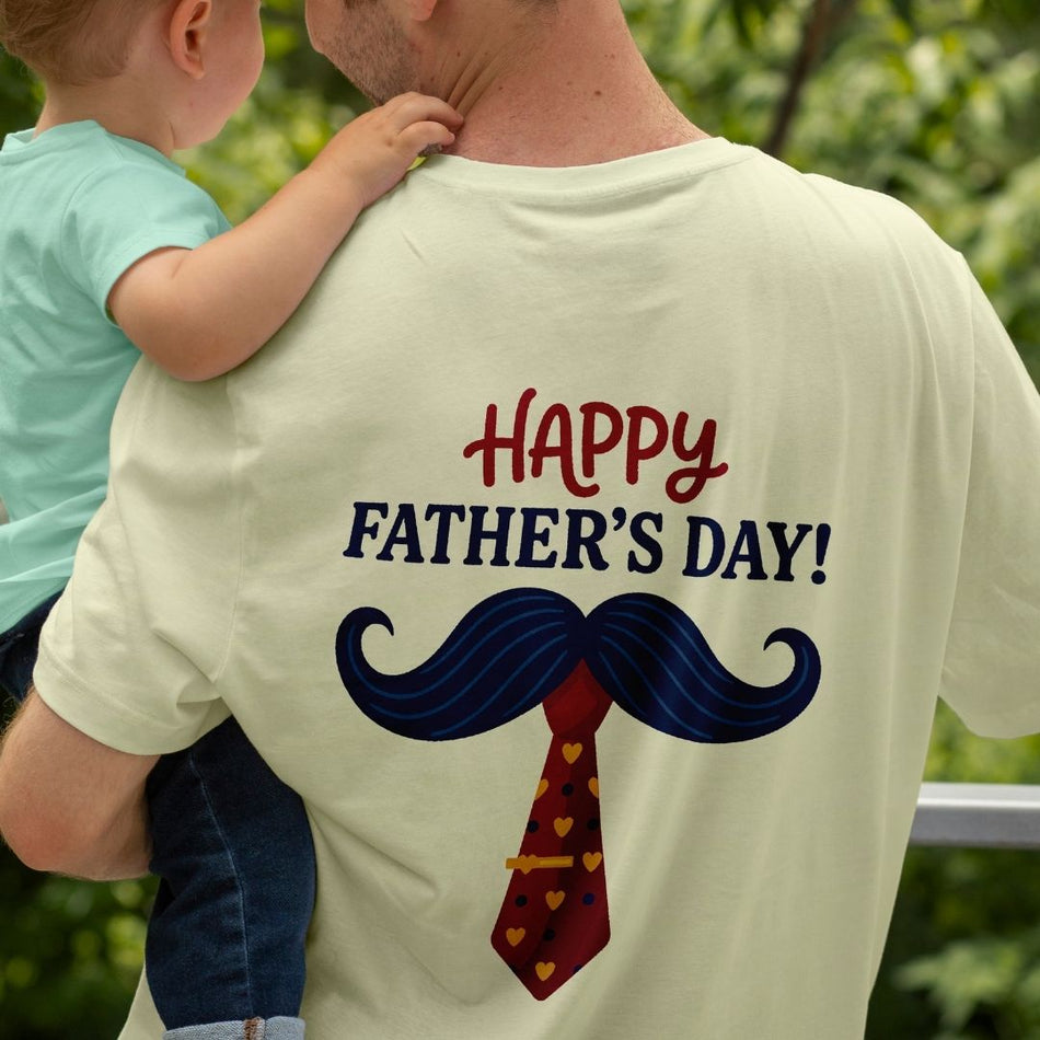 A man wearing a light cream t-shirt with a 'Happy Father's Day!' graphic featuring a blue mustache and red heart-patterned tie, while carrying a child outdoors.