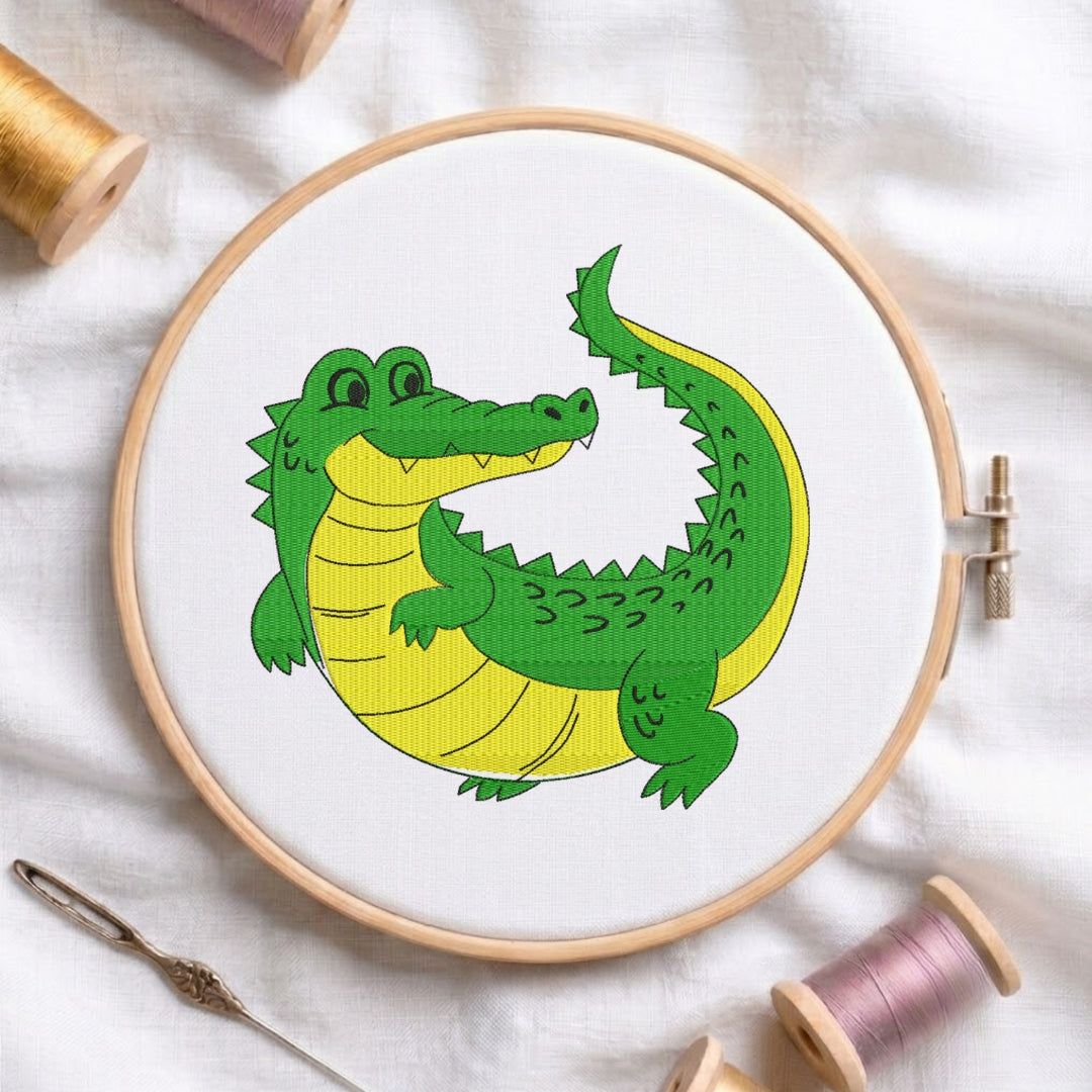 A circular embroidery hoop holding white fabric with a cartoon crocodile design in green and yellow. The hoop is photographed on a white fabric surface with spools of thread and a needle around it.
