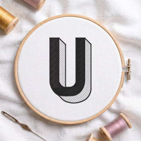 A wooden embroidery hoop holding white fabric with a large black letter U stitched in the center, surrounded by spools of thread and a needle on a white fabric background.
