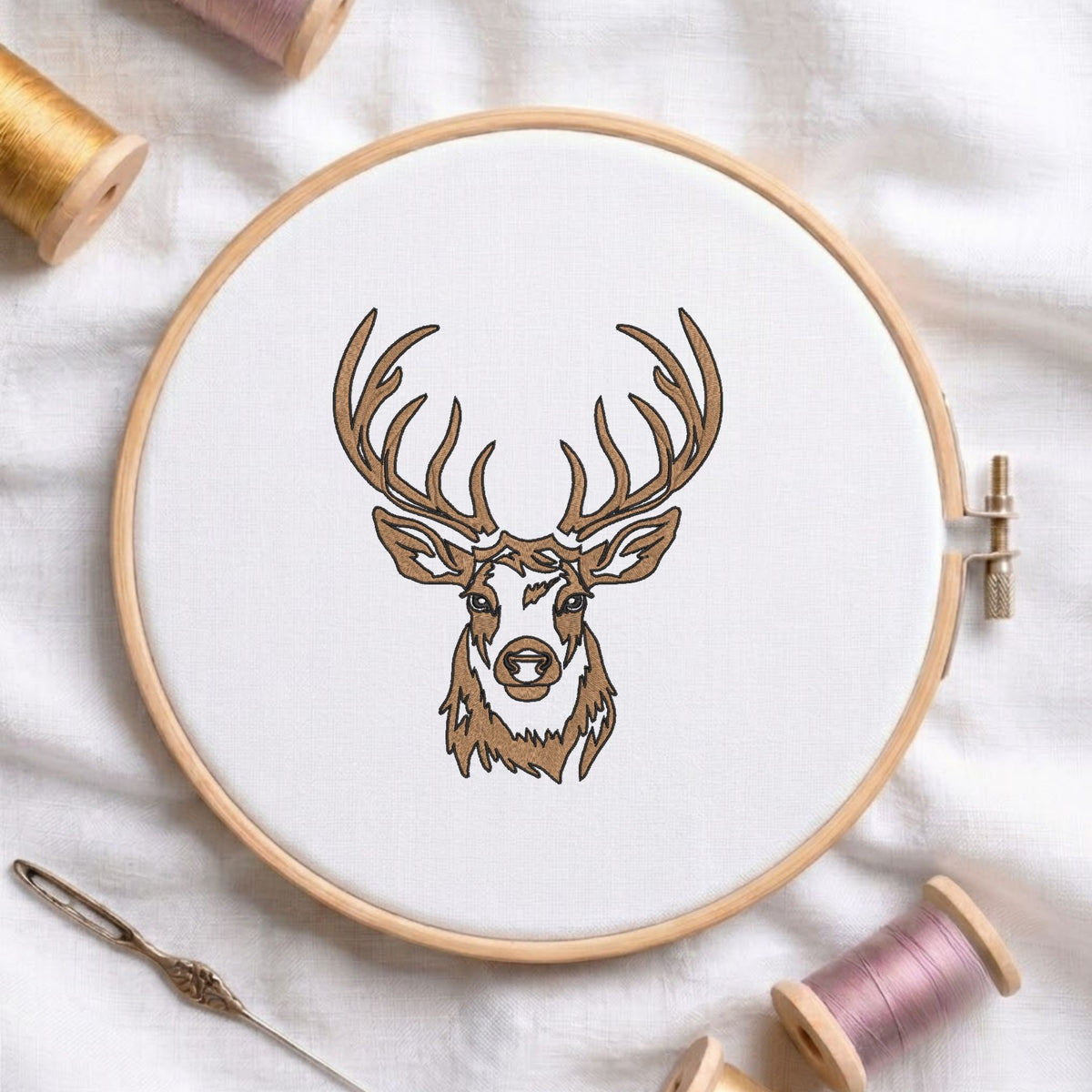 Deer head machine embroidery design displayed in a wooden hoop on white fabric with thread spools and a needle