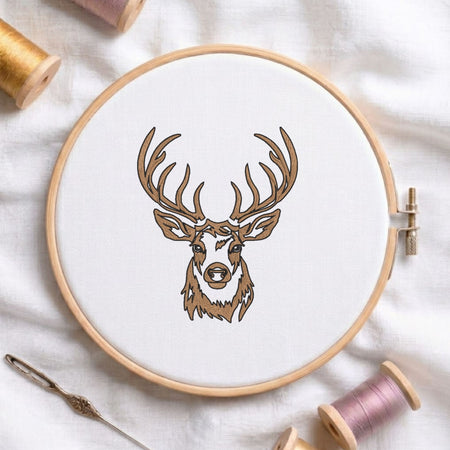 Deer head machine embroidery design displayed in a wooden hoop on white fabric with thread spools and a needle