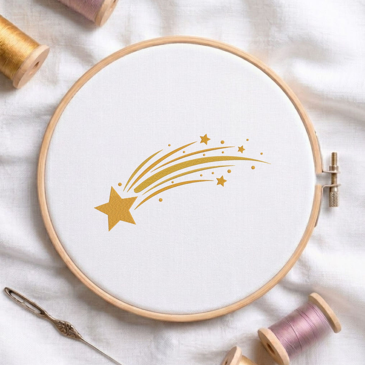 Golden shooting star machine embroidery design displayed in a wooden hoop on white fabric with thread spools and a needle