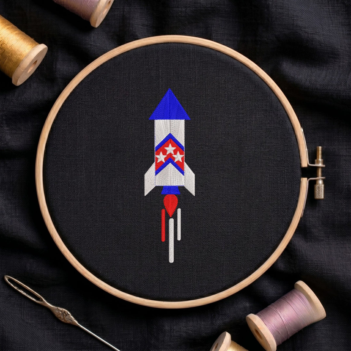Red, white, and blue patriotic space rocket launching with smoke trails machine embroidery design, displayed inside a round wooden hoop on black fabric with thread spools and a needle.