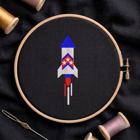 Red, white, and blue patriotic space rocket launching with smoke trails machine embroidery design, displayed inside a round wooden hoop on black fabric with thread spools and a needle.