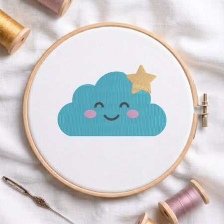 A wooden embroidery hoop holds white fabric with a cute blue cloud design that has a smiling face and a small gold star on top. The hoop is styled on a white fabric background with sewing supplies, including thread spools and a needle.