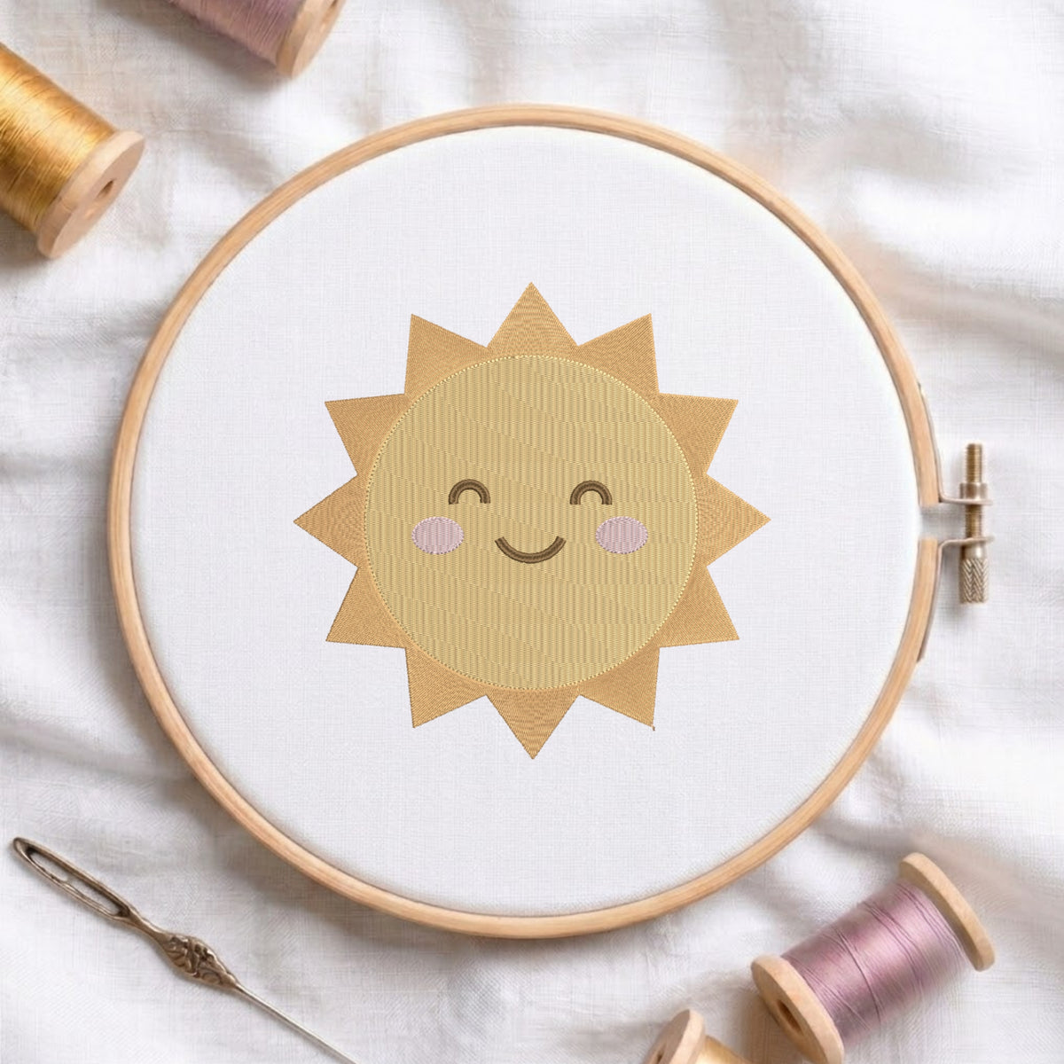 A wooden embroidery hoop holding white fabric with a smiling sun design in tan and light pink thread, styled on a white cloth with spools of thread and a needle around it.