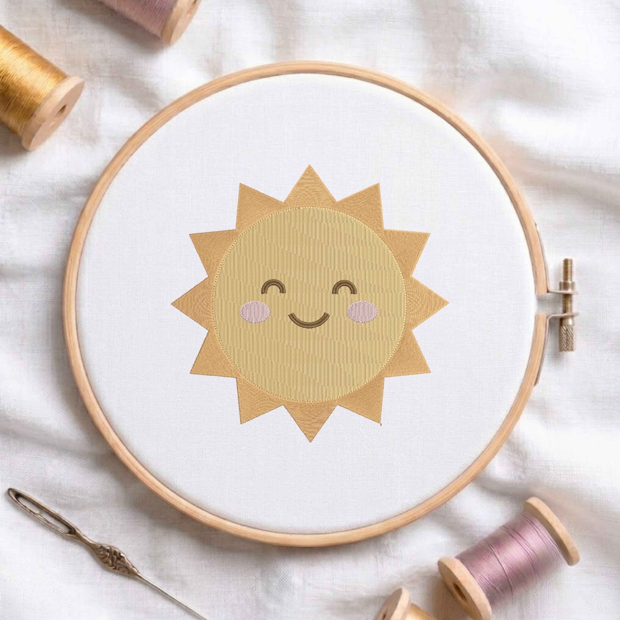 A wooden embroidery hoop holding white fabric with a smiling sun design in tan and light pink thread, styled on a white cloth with spools of thread and a needle around it.