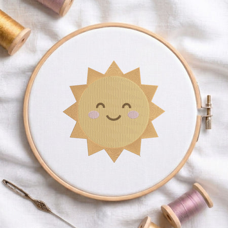 A wooden embroidery hoop holding white fabric with a smiling sun design in tan and light pink thread, styled on a white cloth with spools of thread and a needle around it.