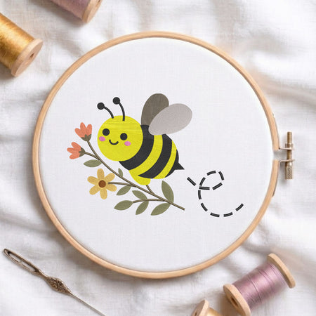 A wooden embroidery hoop holds white fabric with a cute cartoon bee design in yellow, black, gray, pink, and green, surrounded by spools of thread and a needle on a white fabric background.