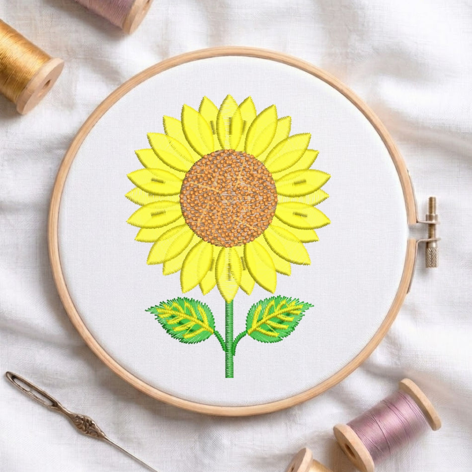 A sunflower embroidery design displayed in a wooden embroidery hoop, surrounded by thread spools and a needle on a white fabric background.