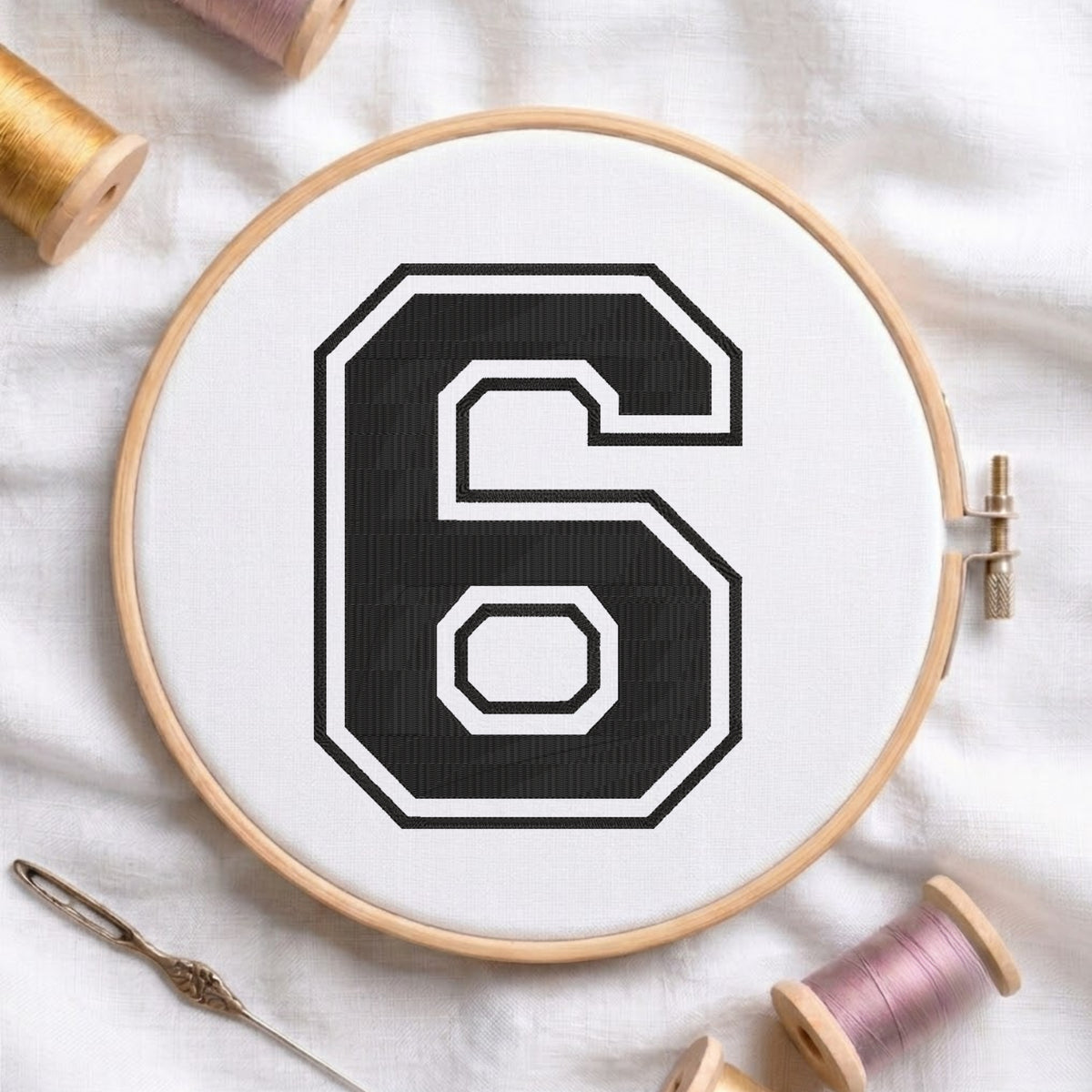 A wooden embroidery hoop holding white fabric with a large black number 6 outlined in white, styled on a white fabric background with spools of thread and a needle.