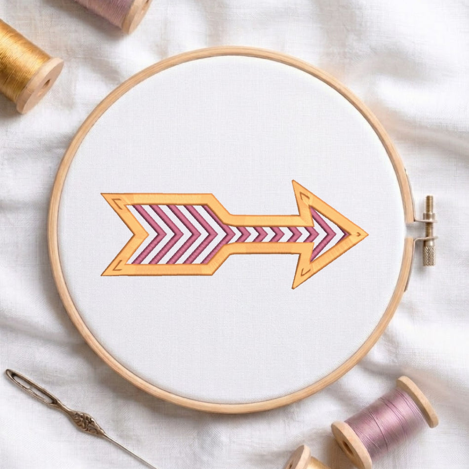 A wooden embroidery hoop holding white fabric, featuring a stylized orange arrow with pink and white chevron stripes. The hoop is styled on a white fabric background with spools of thread and a needle.