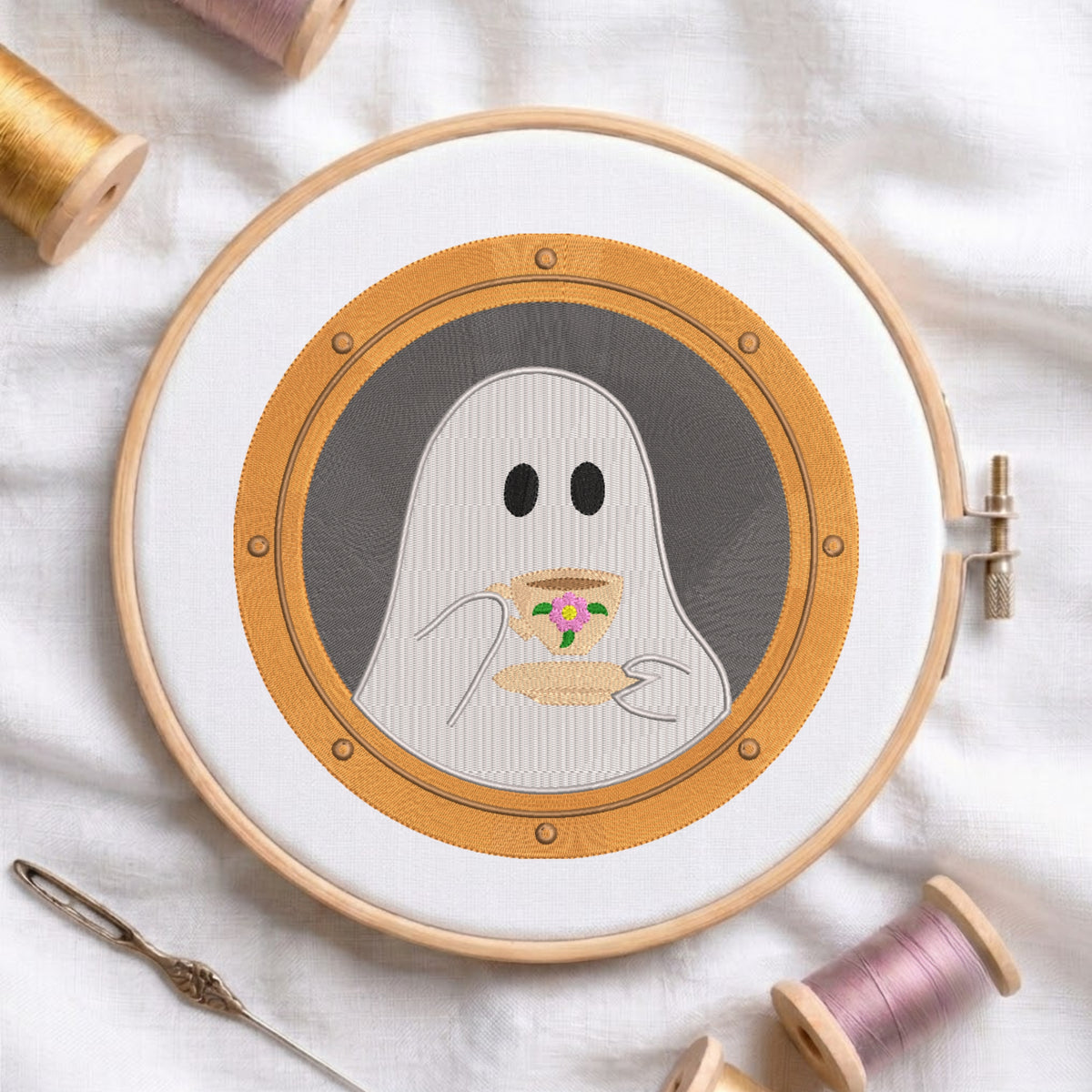 A circular embroidery hoop holding a stitched design of a ghost holding a cup, surrounded by spools of thread and a needle on a white fabric background.