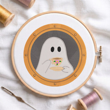 A circular embroidery hoop holding a stitched design of a ghost holding a cup, surrounded by spools of thread and a needle on a white fabric background.