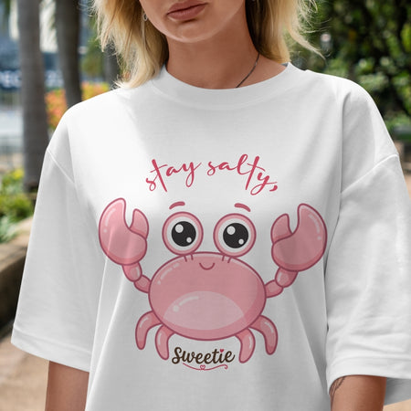White t-shirt with pink crab graphic and text worn by a person outdoors.