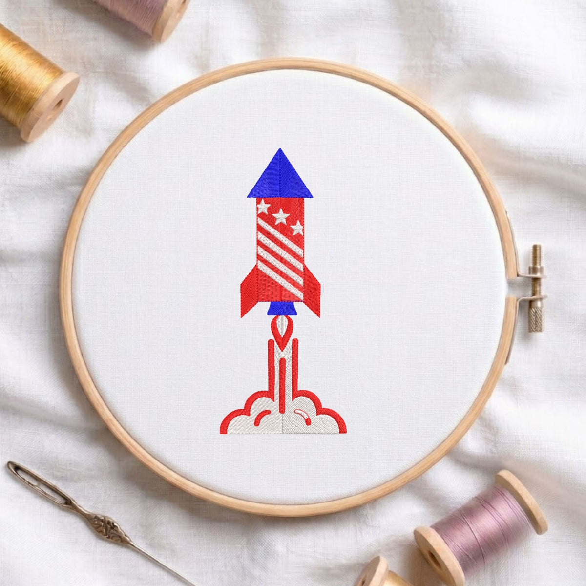 Red, white, and blue patriotic space rocket launching with smoke trails machine embroidery design, displayed inside a round wooden hoop on white fabric with thread spools and a needle