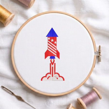 Red, white, and blue patriotic space rocket launching with smoke trails machine embroidery design, displayed inside a round wooden hoop on white fabric with thread spools and a needle