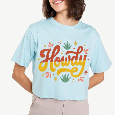 A woman modeling a light pastel blue crop top t-shirt featuring a large retro "Howdy" script text design with mustard yellow and terracotta lettering surrounded by desert leaves.
