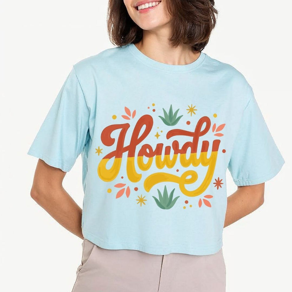 A woman modeling a light pastel blue crop top t-shirt featuring a large retro "Howdy" script text design with mustard yellow and terracotta lettering surrounded by desert leaves.