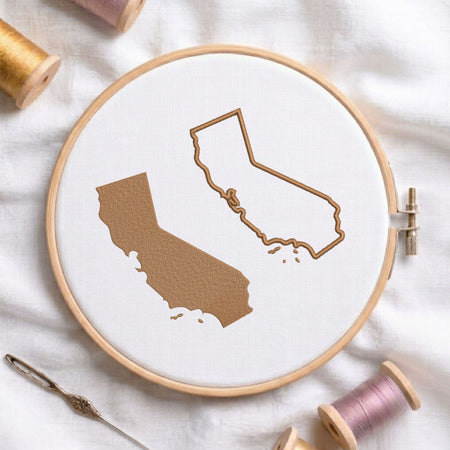 A circular embroidery hoop holding white fabric with two California state outline designs, one filled in tan and one outlined in tan. Spools of thread and a needle are arranged around it on a white fabric background.