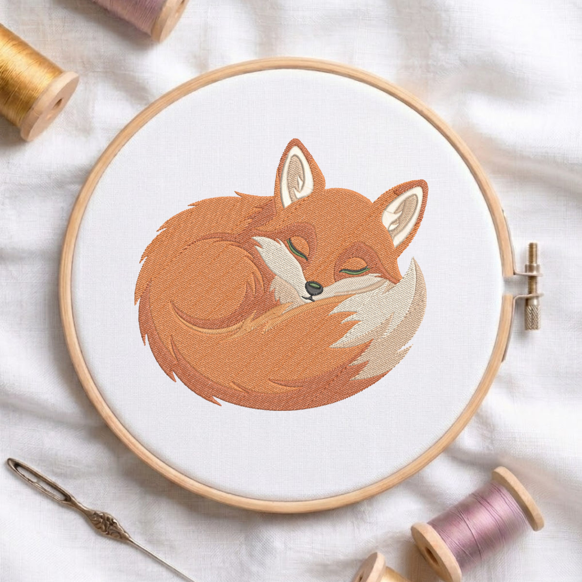 A wooden embroidery hoop on white fabric, featuring a stitched sleeping fox design in warm orange, brown, cream, and olive tones. Spools of thread and a needle are arranged around the hoop in a styled flat-lay composition.