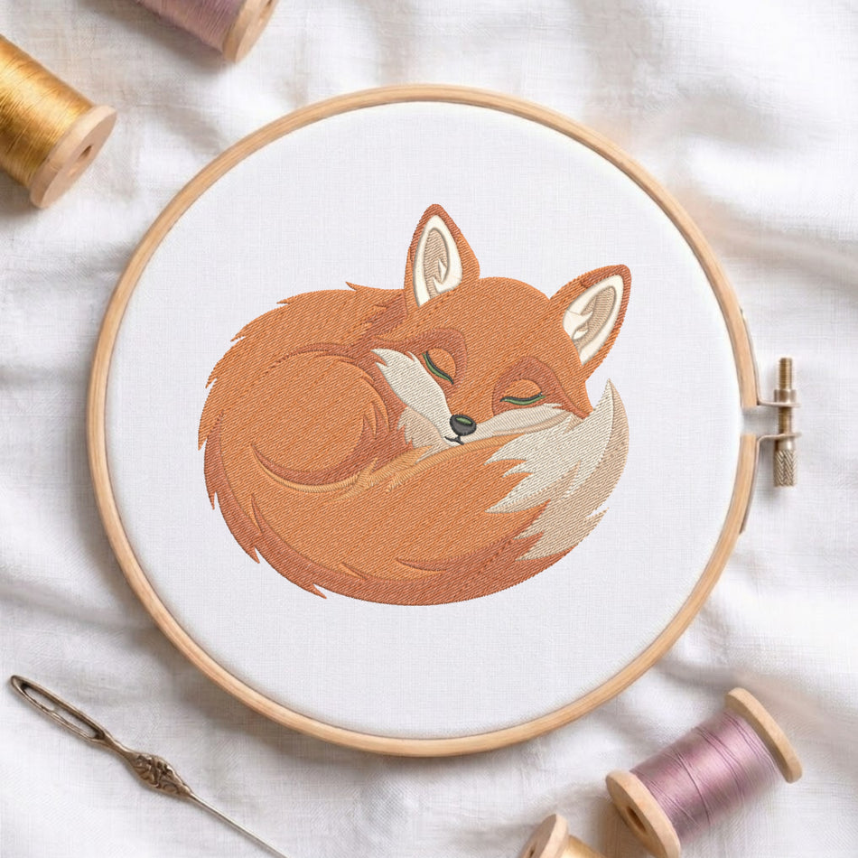 A wooden embroidery hoop on white fabric, featuring a stitched sleeping fox design in warm orange, brown, cream, and olive tones. Spools of thread and a needle are arranged around the hoop in a styled flat-lay composition.