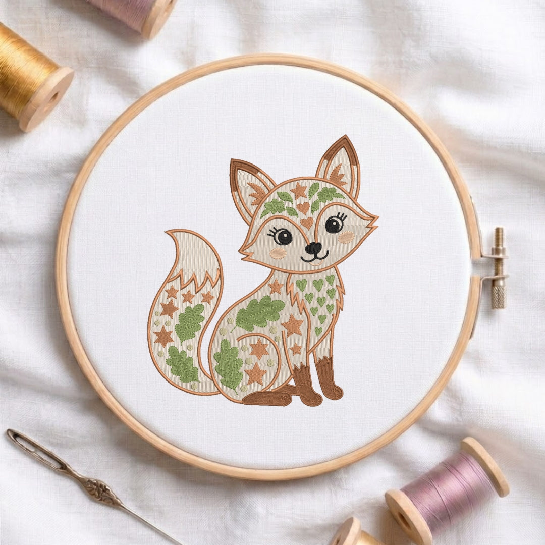 A circular embroidery hoop holding white fabric with a stylized cartoon fox design filled with leaf and star patterns in green, tan, and brown. The hoop is photographed on a white fabric surface with spools of thread and a needle around it.